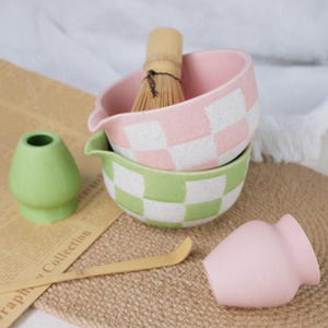 以下が含まれることがあります： 抹茶茶道のセット。ピンクと白のチェック柄のボウル、緑と白のチェック柄のボウル、竹製の泡立て器、抹茶粉末容器、ピンクの茶碗、竹製のスクープが含まれています。茶道に最適です。