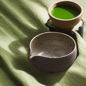 以下が含まれることがあります： 薄い茶色の斑点のある釉薬と濃い茶色の斑点のある釉薬の2つの陶器のボウル。左側のボウルは空で、右側のボウルは明るい緑色の液体が入っています。