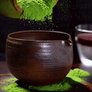 以下が含まれることがあります： 注ぎ口のある濃い茶色の陶器のボウルに、緑色の抹茶パウダーが満たされています。パウダーは木製のさじから注がれています。