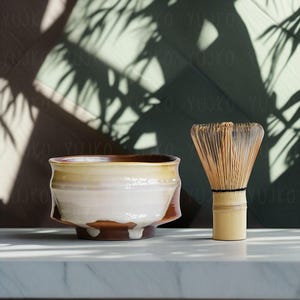 以下が含まれることがあります： 白いセラミックの抹茶ボウルに茶色の縁取り、竹製の抹茶ホイスクが置かれた白い大理石の表面