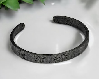 May include: A dark gray cuff bracelet with a textured, wood-grain pattern. The open-ended bracelet is made of metal and has a circular design. The bracelet is approximately 2.5 inches in diameter.