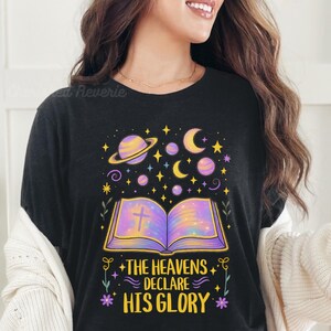 Op de afbeelding: Zwart T-shirt met een hemels ontwerp met een open boek met een kruis, planeten, sterren en manen in paars, geel en blauw. De tekst luidt "THE HEAVENS DECLARE HIS GLORY".