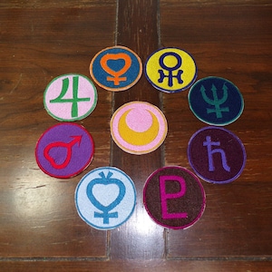 May include: Nine round embroidered patches with different colors and symbols, including a crescent moon, a heart, a star, and a trident. The patches are arranged in a circle on a wooden surface.