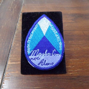 May include: A blue, white, and teal embroidered patch in the shape of a teardrop. The patch says "Maybe I'm not Alone" and "Distant Shore" on the sides.