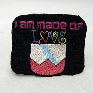 May include: Black embroidered patch with the text "I am made of love" in pink and white lettering. The patch features a pink and white heart with a rainbow design.