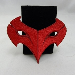 May include: A vibrant red embroidered mask with a pointed bottom and eye cutouts. The mask is displayed on a black surface against a white background. The mask's design is symmetrical and detailed.