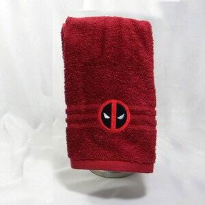 May include: A red bath towel with a white embroidered Deadpool logo. The logo is a red circle with a black outline and a white mask with black eyes.
