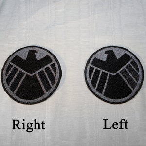 May include: Two black and gray embroidered patches with the S.H.I.E.L.D. logo. The logo is a black eagle with a shield in front of it. The patches are on a white background. The word "Right" is below the patch on the right and the word "Left" is below the patch on the left.