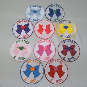 May include: Ten round embroidered patches with a white background, a pink, blue, or yellow bow with a heart in the centre, and the text "FIGHT LIKE A GIRL".