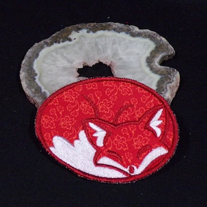 May include: A red fabric patch with a white embroidered sleeping fox design. The fox is curled up with its tail over its nose. The patch has a floral pattern.