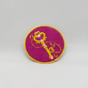 May include: A round pink fabric patch with a yellow embroidered key design. The key has a heart-shaped head and is surrounded by floral details.