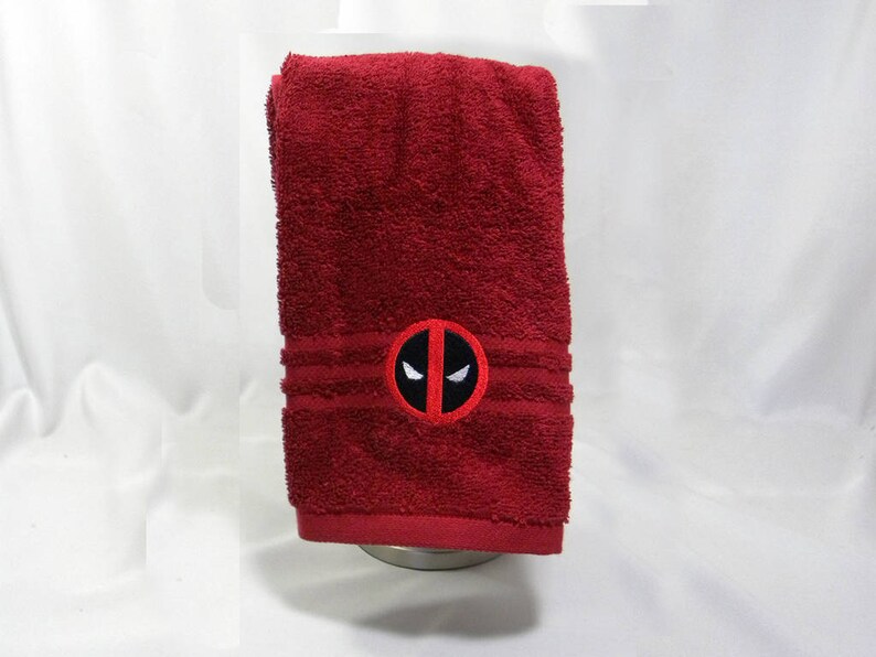 May include: A red bath towel with a white embroidered Deadpool logo. The logo is a red circle with a black outline and a white mask with black eyes.