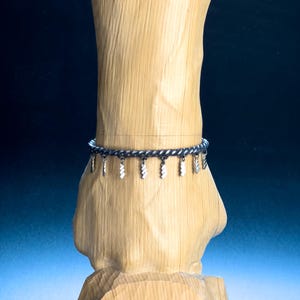 Op de afbeelding: Een zwarte gevlochten armband met zilverkleurige bedels die eraan hangen. De armband wordt getoond op een houten oppervlak.
