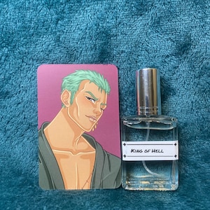 Pode incluir: Um frasco de perfume com tampa prateada e um rótulo branco que diz "KING OF HELL" ao lado de um cartão com uma ilustração de desenho animado de uma pessoa com cabelo verde. O fundo é um tecido texturizado azul-petróleo.