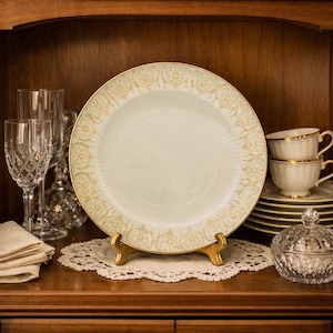 May include: A decorative plate with a gold rim and ornate gold detailing, displayed on a gold stand. The plate is light green with a gold floral pattern. Crystal glassware, teacups, and a sugar bowl are also visible.