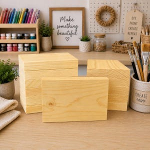 May include: Several unfinished wooden blocks are stacked and arranged on a light-colored wooden surface. The blocks are rectangular and have a natural wood grain. The background includes art supplies, a sign that says "Make something beautiful", and a container of paint brushes.