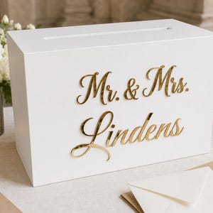 Könnte beinhalten: Eine rechteckige weiße Kartenbox mit einem horizontalen Schlitz an der Oberseite und goldfarbenen, reflektierenden Schreibschriftbuchstaben auf der Vorderseite, die Mr. & Mrs. Lindens lauten. Die Box hat eine glatte Oberfläche.