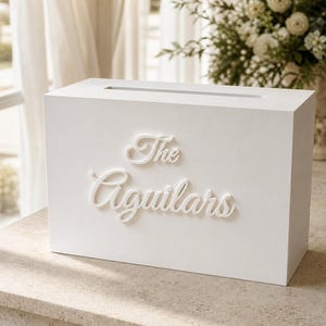 Könnte beinhalten: Eine rechteckige weiße Holzbox mit einem horizontalen Schlitz auf der Oberseite. Die Vorderseite der Box ist mit erhabenen weißen Buchstaben in Schreibschrift versehen, die The Aguilars lauten. Die Box hat eine glatte, matte Oberfläche.