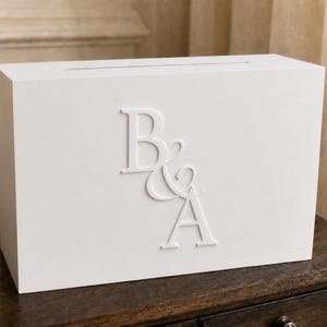 May include: A rectangular white wooden box with a horizontal slot on the top surface. The front panel features raised, three-dimensional white lettering displaying the characters B & A in a serif font style, centered on the smooth white surface.