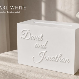 Puede incluir: Una caja para tarjetas rectangular de color blanco perla con una ranura horizontal en la parte superior. El panel frontal muestra el texto blanco en relieve "Danet and Jonathan" en letra cursiva. La caja tiene un acabado liso y sólido.