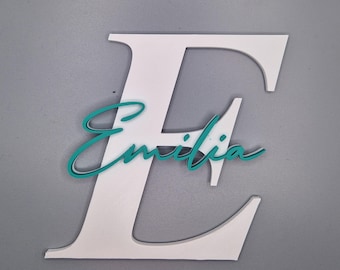 Puede incluir: Una letra "E" de madera blanca con el nombre "Emilia" en escritura turquesa superpuesto. El nombre está ligeramente desplazado, creando un efecto de sombra. El fondo es gris liso.