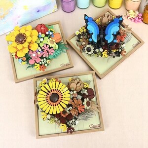 Pode incluir: Três molduras quadradas de madeira com fundo castanho e moldura branca. Cada moldura contém um arranjo floral 3D feito de peças de plástico coloridas. Uma moldura apresenta uma borboleta azul, outra apresenta um girassol amarelo e a terceira apresenta um girassol amarelo com detalhes castanhos.