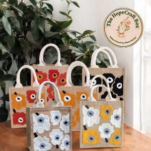 Op de afbeelding: Zes draagtassen gemaakt van natuurlijke jute stof met witte handvatten. Elke tas heeft een ander geborduurd bloemenmotief in verschillende kleuren, waaronder oranje, geel, wit en zwart.