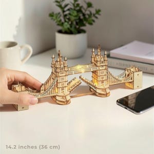 Może przedstawiać: Szczegółowy drewniany model Tower Bridge, o długości 36 cm, z wewnętrznym oświetleniem. Model z jasnego drewna przedstawia wieże i konstrukcję wiszącą mostu. Model jest umieszczony na białej powierzchni, widoczny jest smartfon.