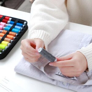 May include: A person is holding a spool of gray thread and sewing a white shirt. The thread is in a plastic container with a white label. The person is using their right hand to hold the thread and their left hand to hold the shirt.