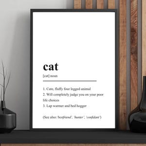 Peut inclure: Une affiche encadrée avec la définition du mot "chat" en texte noir sur fond blanc. La définition comprend trois points sur les chats, et l'affiche est exposée sur une étagère noire avec des objets décoratifs.