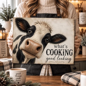May include: A rectangular sign featuring a black and white cow illustration with the text "what's COOKING good looking" in black lettering. The sign has a light, weathered background and a small heart icon below the text.