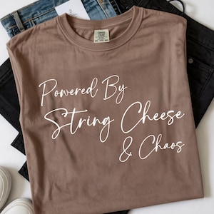 Op de afbeelding: Een bruin T-shirt met de tekst "Powered By String Cheese & Chaos" in witte letters. Het shirt is opgevouwen, met blauwe en zwarte jeans op de achtergrond. Het shirt is maat XXL.