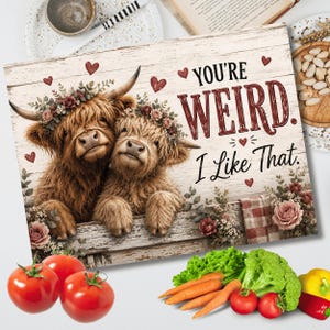 May include: A rectangular sign featuring two brown Highland cows with floral crowns on a white wood-grain background. The text reads "YOU'RE WEIRD. I Like That." surrounded by small red hearts and floral accents at the bottom corners.