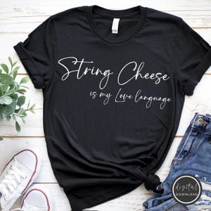 Op de afbeelding: Zwart T-shirt met de witte tekst "String Cheese is my Love Language" in een sierlijk lettertype. Het shirt is gestyled met opgerolde mouwen en een knoop aan de onderkant. Witte sneakers en een blauwe jeans zijn zichtbaar.