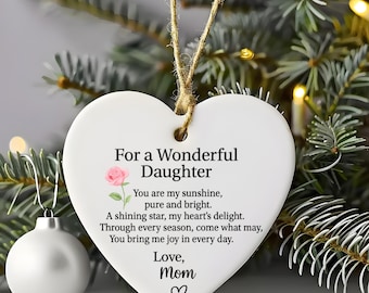 May include: A white heart-shaped ceramic ornament hanging from a jute string. It features a pink rose illustration and black text that reads: For a Wonderful Daughter, You are my sunshine, pure and bright. A shining star, my heart's delight.