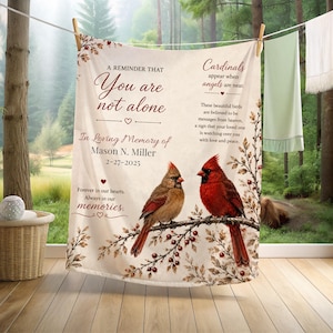 May include: A cream-colored blanket with a memorial message and cardinal birds. The blanket features the text "You are not alone" and "In Loving Memory of Mason N. Miller 2-27-2025". Two cardinals are perched on a branch with berries.