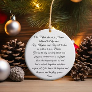 Peut inclure: Ornement de Noël en céramique blanche avec la prière du Notre Père imprimée en script noir. L'ornement est suspendu à une ficelle de ficelle naturelle, entouré de pommes de pin et de décorations de Noël.