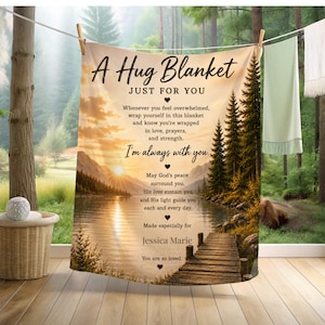 May include: A beige blanket with the text "A Hug Blanket Just For You" and a scenic lake and mountain design. The blanket includes the text "I'm always with you" and other inspirational text. The blanket is hanging on a clothesline.