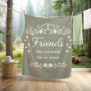 Peut inclure: Une couverture rectangulaire vert sauge suspendue à une corde à linge, arborant un motif imprimé blanc composé de deux cœurs, de vignes fleuries et du texte "Friends like you make life so sweet" centré, entouré de petites icônes de cœurs.