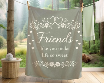 May include: A sage green rectangular blanket hanging on a clothesline, featuring a white printed design of two hearts, floral vines, and the text "Friends like you make life so sweet" centered on the fabric, surrounded by small heart icons.