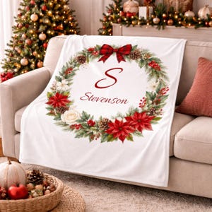 May include: White Christmas blanket with a wreath design. The wreath includes red poinsettias, pinecones, and a plaid bow. The letter "S" and the name "Stevenson" are in the centre. The blanket is on a beige sofa.