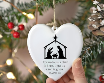 May include: White heart-shaped Christmas ornament with a black silhouette of the Nativity scene and the text "For unto us a child is born, unto us a son is given. ISAIAH 9:6". The ornament is hanging from a twine string.