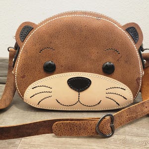 May include: A brown, semi-circular crossbody bag shaped like an otter face. It features a lighter beige muzzle, black stitched whiskers, a black nose, and two round black eyes. The bag has small ears at the top and a long, adjustable brown strap.