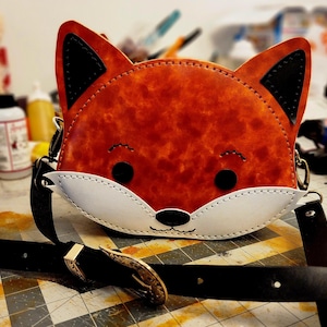 May include: A fox-shaped crossbody bag crafted from orange leather with a white muzzle, black ears, and black circular eyes. The bag features a black leather strap with an ornate metal buckle and is stitched with white thread along the edges.