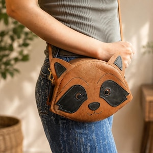 May include: A tan leather crossbody bag shaped like a raccoon face, featuring dark brown leather patches around the eyes, pointed ears, a small dark nose, and stitched details. The bag includes a thin leather shoulder strap and gold hardware.