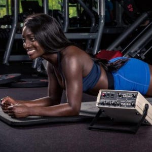 Op de afbeelding: Een vrouw in blauwe sportkleding doet een plankoefening op een mat. Een beige elektronisch apparaat met knoppen en schakelaars staat voor haar. Op de achtergrond is fitnessapparatuur te zien.