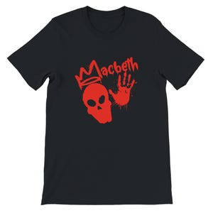 Peut inclure: T-shirt noir avec un motif rouge. Le motif comprend le mot "Macbeth", une tête de mort couronnée et une empreinte de main. Le graphisme a un aspect vieilli, comme peint à la bombe. Le t-shirt est à manches courtes.