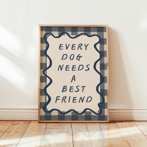 Op de afbeelding: Een ingelijste print met de tekst "EVERY DOG NEEDS A BEST FRIEND". De print heeft een crèmekleurige achtergrond met de tekst in donkerblauw. De lijst is licht van kleur en de achtergrond is een blauw-wit geblokt patroon.