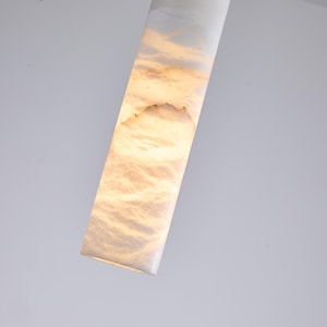 Puede incluir: Una luminaria rectangular e iluminada, con un exterior blanco y un interior cálido en forma de nube. La luminaria está en ángulo diagonalmente contra un fondo blanco, mostrando su superficie texturizada y su suave brillo.