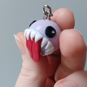 May include: A novelty keyring with a monster head design. The head is a pale purple colour, with black eyes, white teeth, and a red tongue. A silver-coloured metal ring is attached.